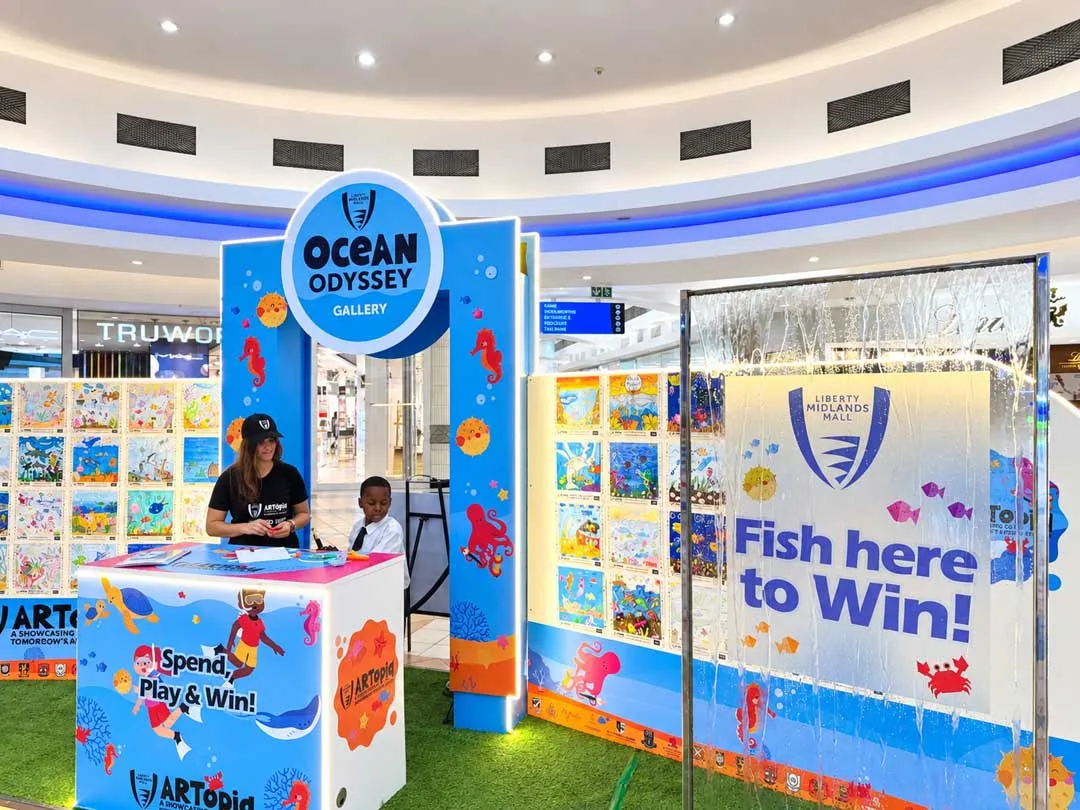 The ARTopia Ocean Odyssey Gallery displays children's artwork beside a Fish Here to Win promotional installation at Liberty Midlands Mall.